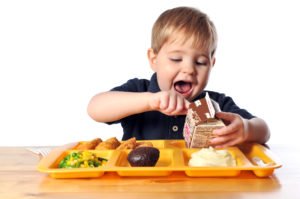 Image of young boy who is sitting down to eat his cafeteria school lunch. He has chicken, mashed potatoes, mixed vegetables, brownie and chocolate milk. He is inspecting the milk carton and likes what he sees.