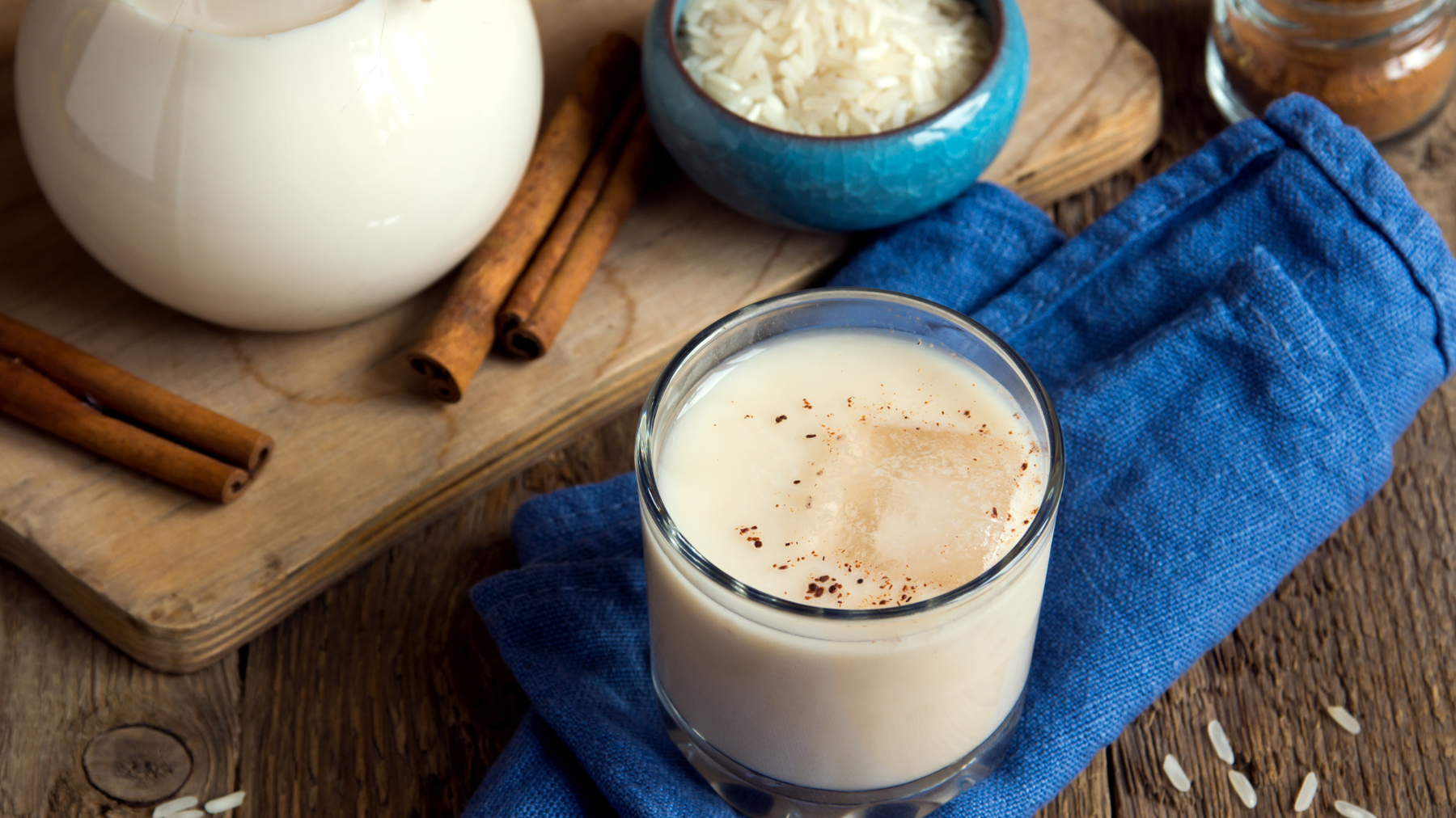 A glass of horchata over ice displayed on a blue napkin with a bowl of dried rice and pitcher of milk.